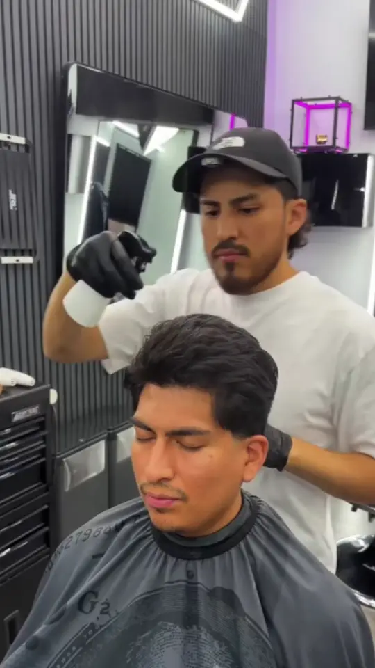 Alex, in a black cap and gloves, fading the side of his brother's hair in the shop